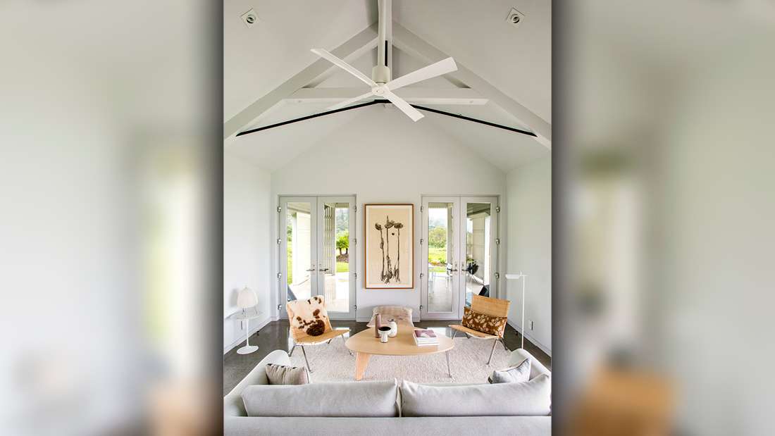 Bright modern living room with vaulted ceiling featuring exposed beams and white ceiling fan, minimalist decor with light gray sofa, wooden coffee table, cowhide accent chair, and double French doors opening to outdoor patio.