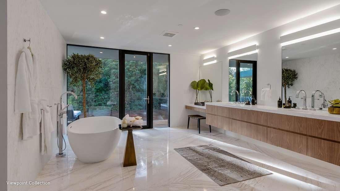 Luxury modern bathroom with freestanding oval soaking tub, marble flooring and walls, double vanity with wood cabinetry, large mirrors, and floor-to-ceiling glass doors opening to a private outdoor deck.