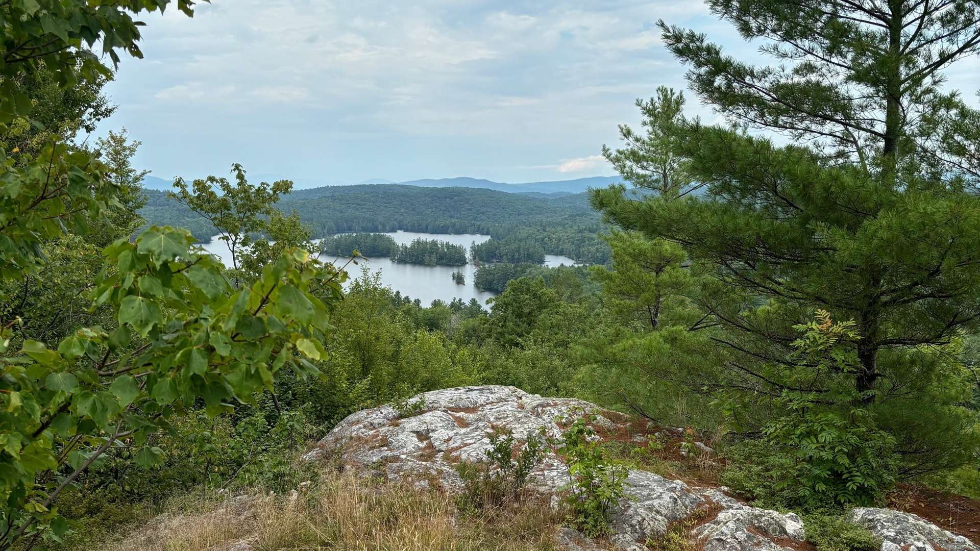 Rocky hilltop with green trees overlooking a lake dotted with forested islands under a partly cloudy sky.