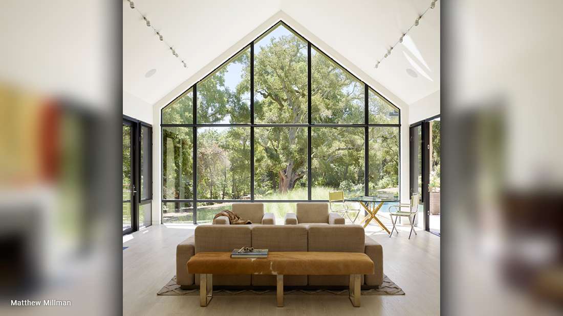 Modern living room with vaulted ceiling and full-height triangular glass wall offering lush garden views, beige sofa with matching ottoman, tan bench, and minimalist decor with light wood flooring.