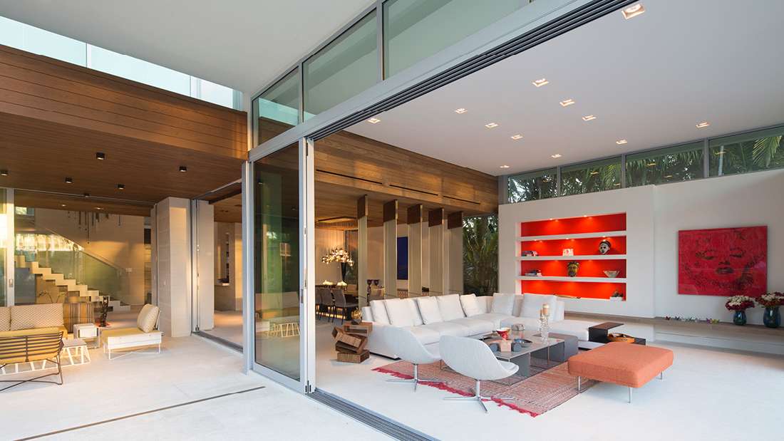 Modern open-concept living room with large sliding glass doors, white sectional sofa, contemporary lounge chairs, and vibrant red built-in shelving, featuring wood ceiling accents and seamless indoor-outdoor design.
