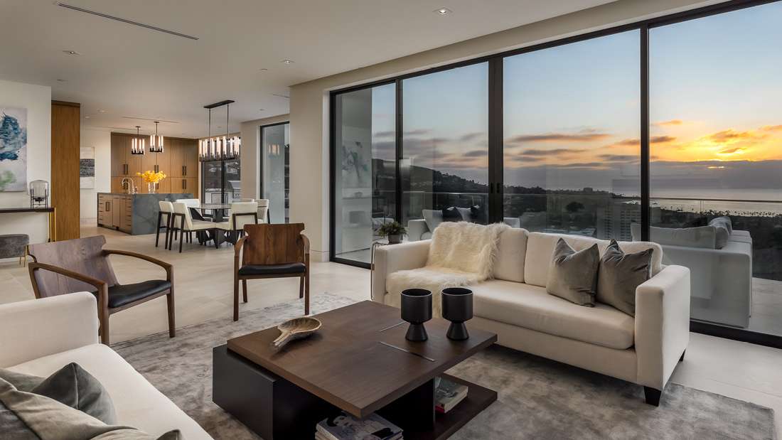 Elegant open-concept living and dining area with modern furniture, floor-to-ceiling glass doors, and breathtaking sunset views over the ocean.