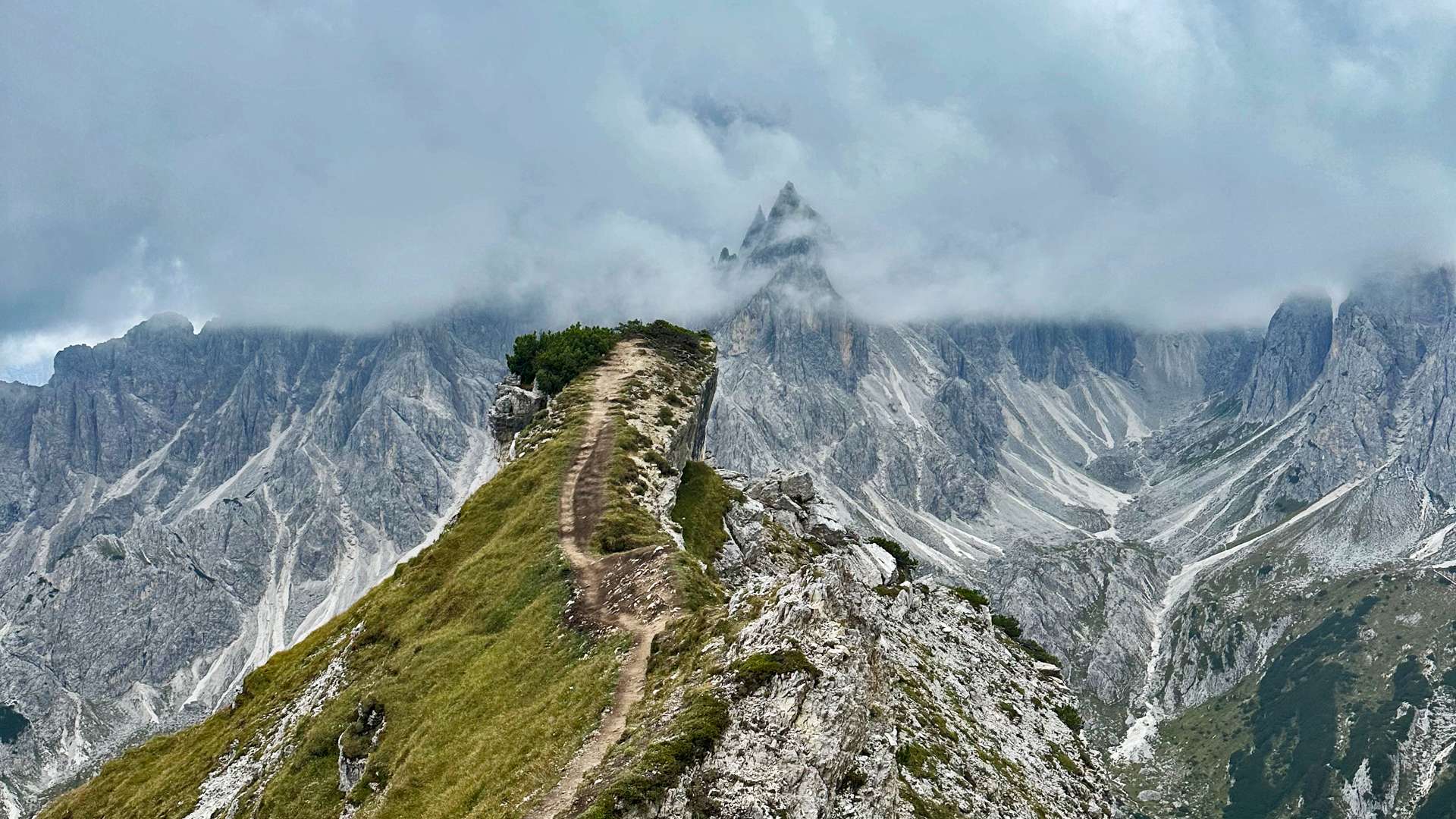 Narrow mountain ridge trail with green grass and rocky slopes leading toward jagged peaks partially hidden by thick clouds.