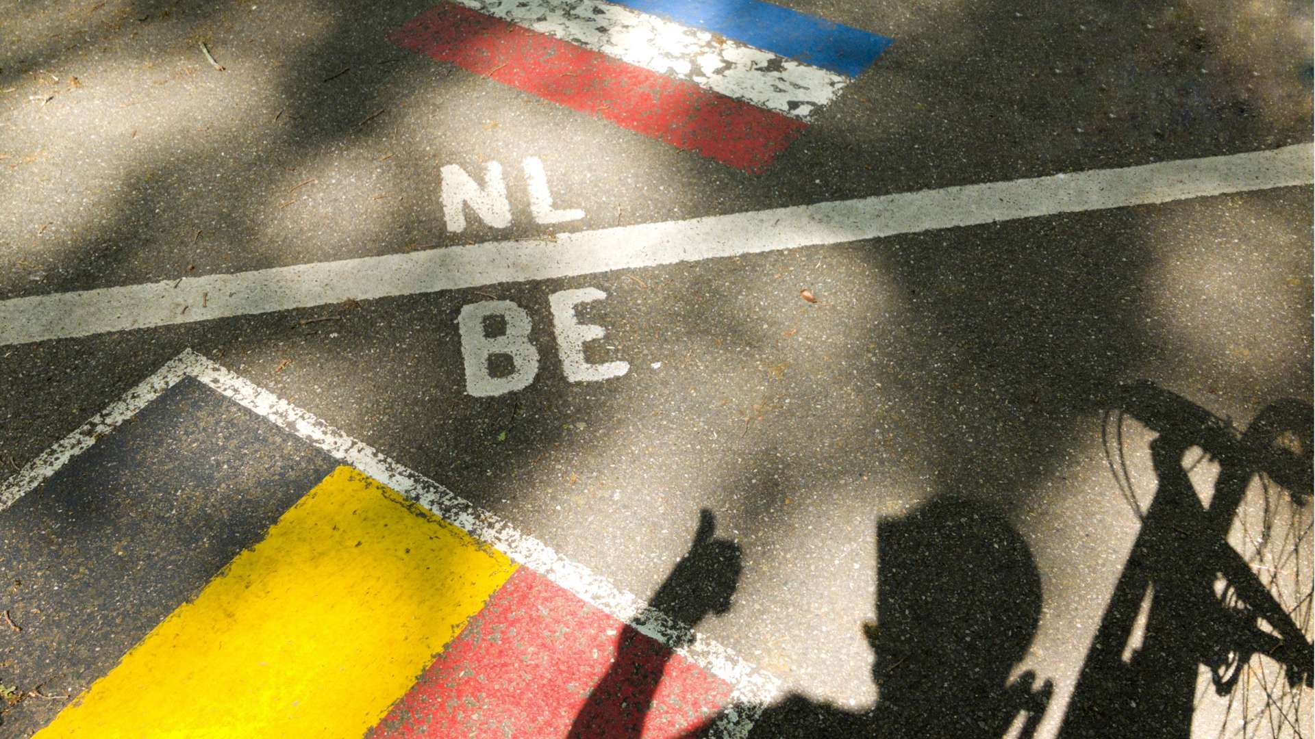 Painted border line on pavement marking the boundary between the Netherlands (NL) and Belgium (BE), with national flag colors on each side and a bicycle shadow in the foreground.