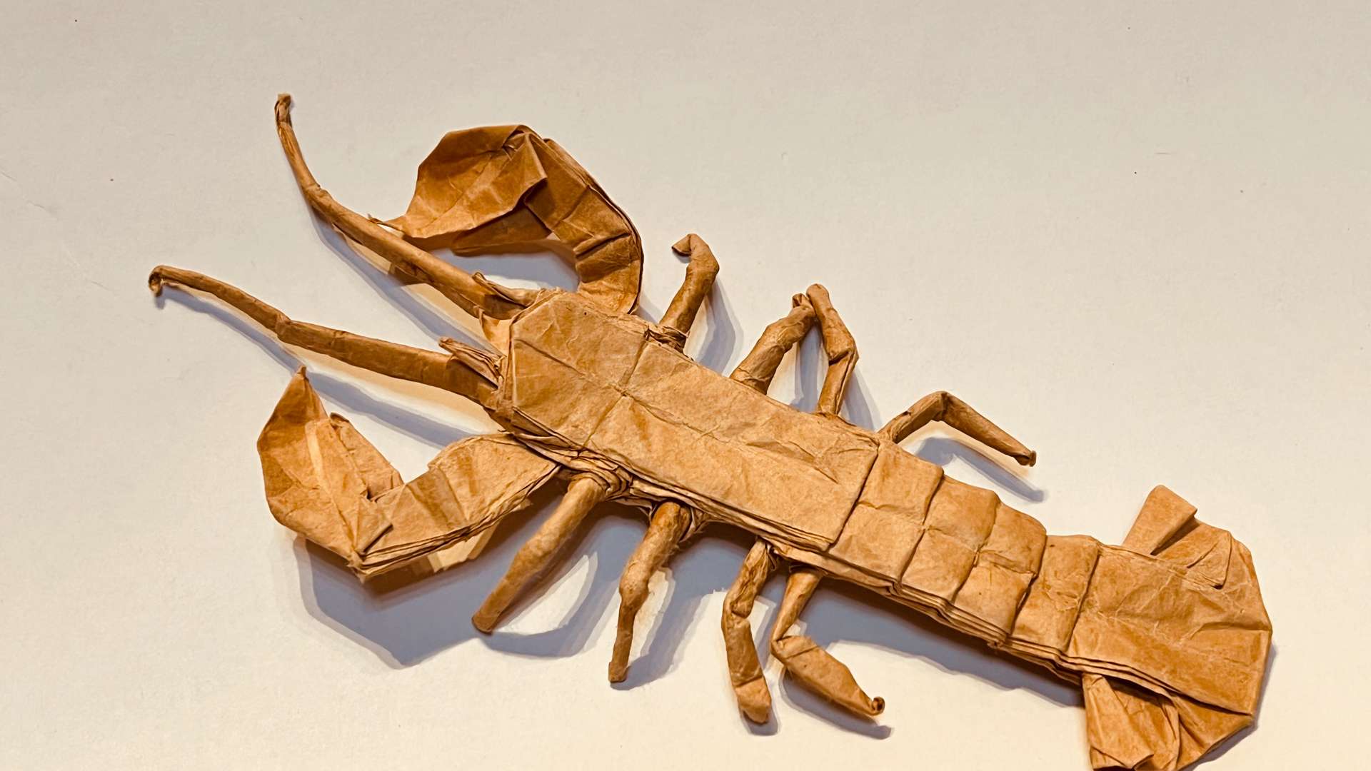   Origami lobster crafted from brown paper with detailed claws, legs, and tail on a light background.