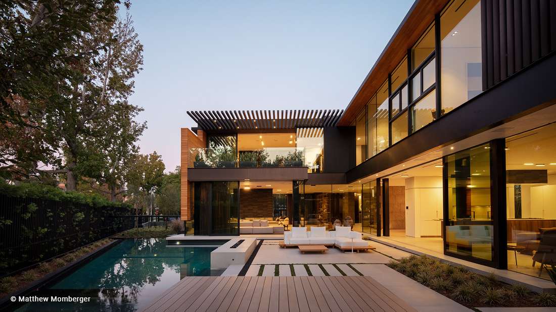 Contemporary luxury home with two-story glass walls, outdoor lounge area, sleek swimming pool, and modern pergola accents surrounded by lush greenery.