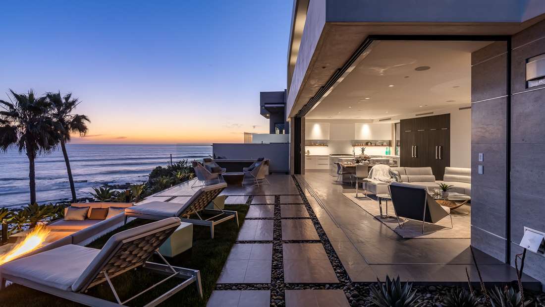  Luxury oceanfront home with open-concept living area, modern kitchen, and outdoor terrace featuring lounge chairs and fire pit overlooking sunset views of the Pacific.