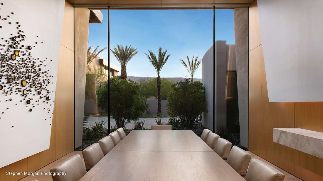 Modern dining room with large floor-to-ceiling glass windows, natural wood table, upholstered chairs, and view of palm trees and contemporary architecture outside.