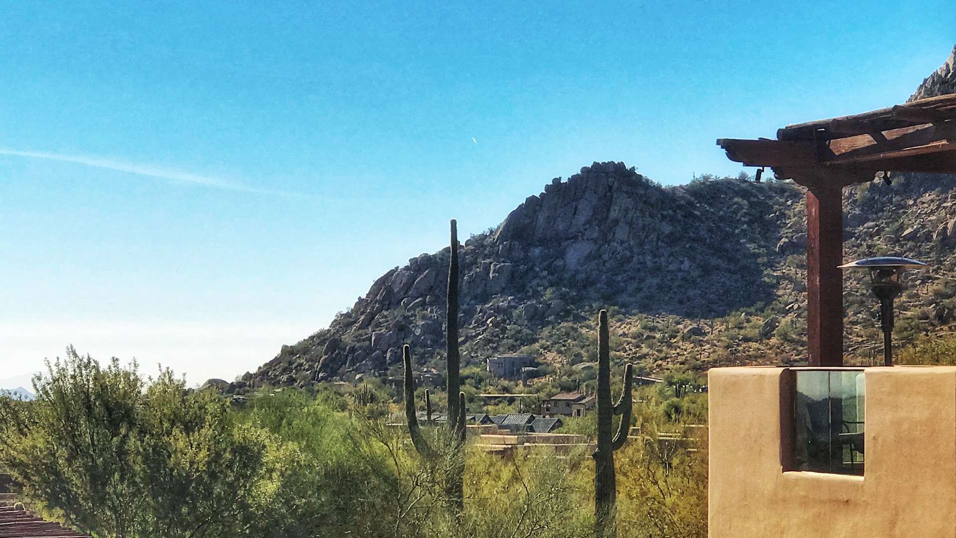   Desert landscape with tall cacti, rocky mountain backdrop, and adobe-style structure under a clear blue sky.
