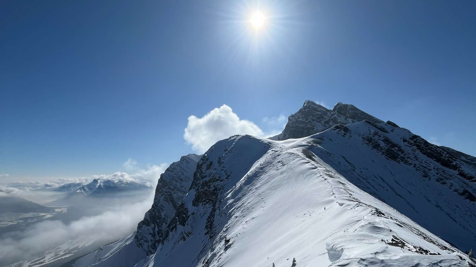Snow-covered mountain ridge under bright sunlight with clear blue sky and scattered clouds above a valley.