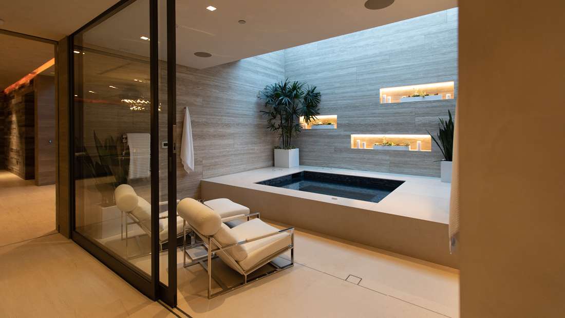 Luxury indoor spa with modern soaking tub, wood-paneled walls, recessed shelving with ambient lighting, potted plants, and glass doors opening to a relaxation area.