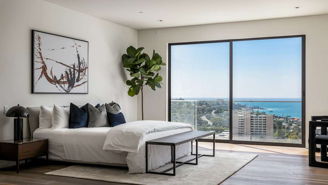Modern bedroom with minimalist decor, white bedding, abstract wall art, and floor-to-ceiling glass doors showcasing panoramic ocean views.