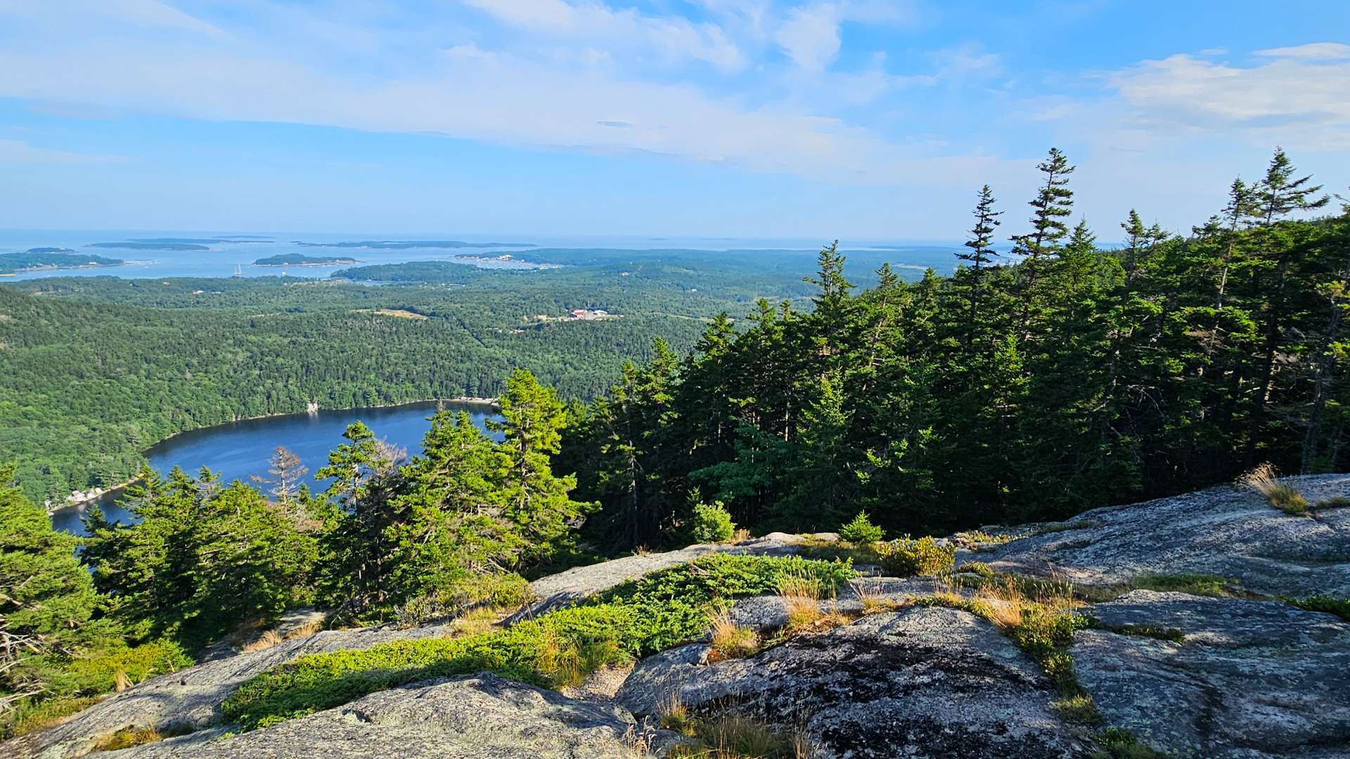 View from rocky mountaintop overlooking dense forest, a blue lake, and distant islands under a clear sky.