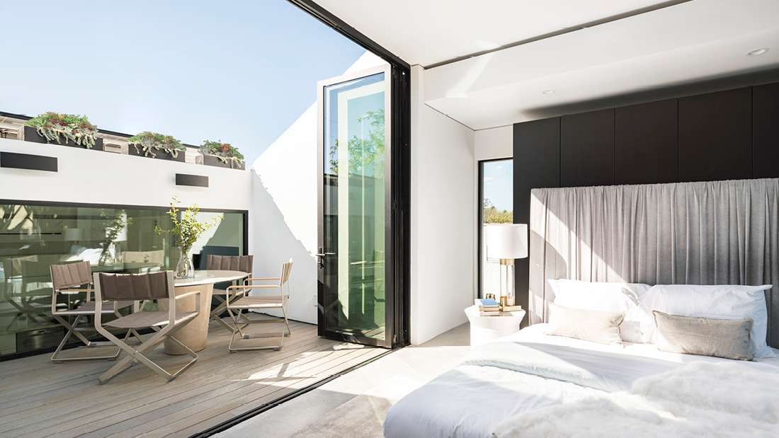 Modern bedroom with white bedding and gray upholstered headboard, folding glass doors opening to a private terrace with outdoor dining set, light wood flooring, and minimalist decor.