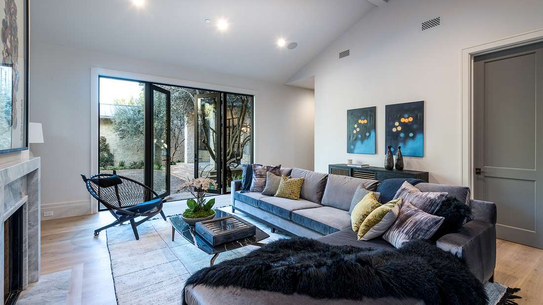 Modern living room with folding glass doors opening to a courtyard, gray sectional sofa with colorful accent pillows, black woven lounge chair, marble fireplace, and minimalist decor featuring abstract wall art.