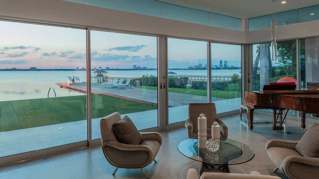 Modern waterfront living room with floor-to-ceiling sliding glass doors offering panoramic views of Biscayne Bay, featuring beige armchairs around a glass coffee table and a polished wooden grand piano.
