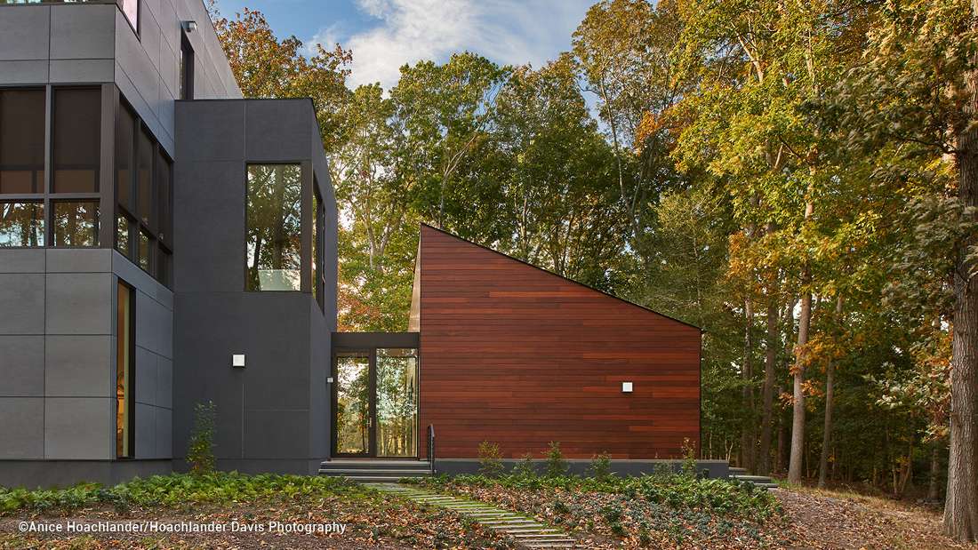 Contemporary home exterior with dark gray panel siding and contrasting sloped section clad in warm wood, large black-framed windows, and glass entry door surrounded by lush forest landscape.”