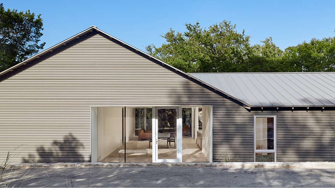 Contemporary single-story home with corrugated metal siding, pitched roof, and large sliding glass doors opening to a minimalist interior with neutral tones.