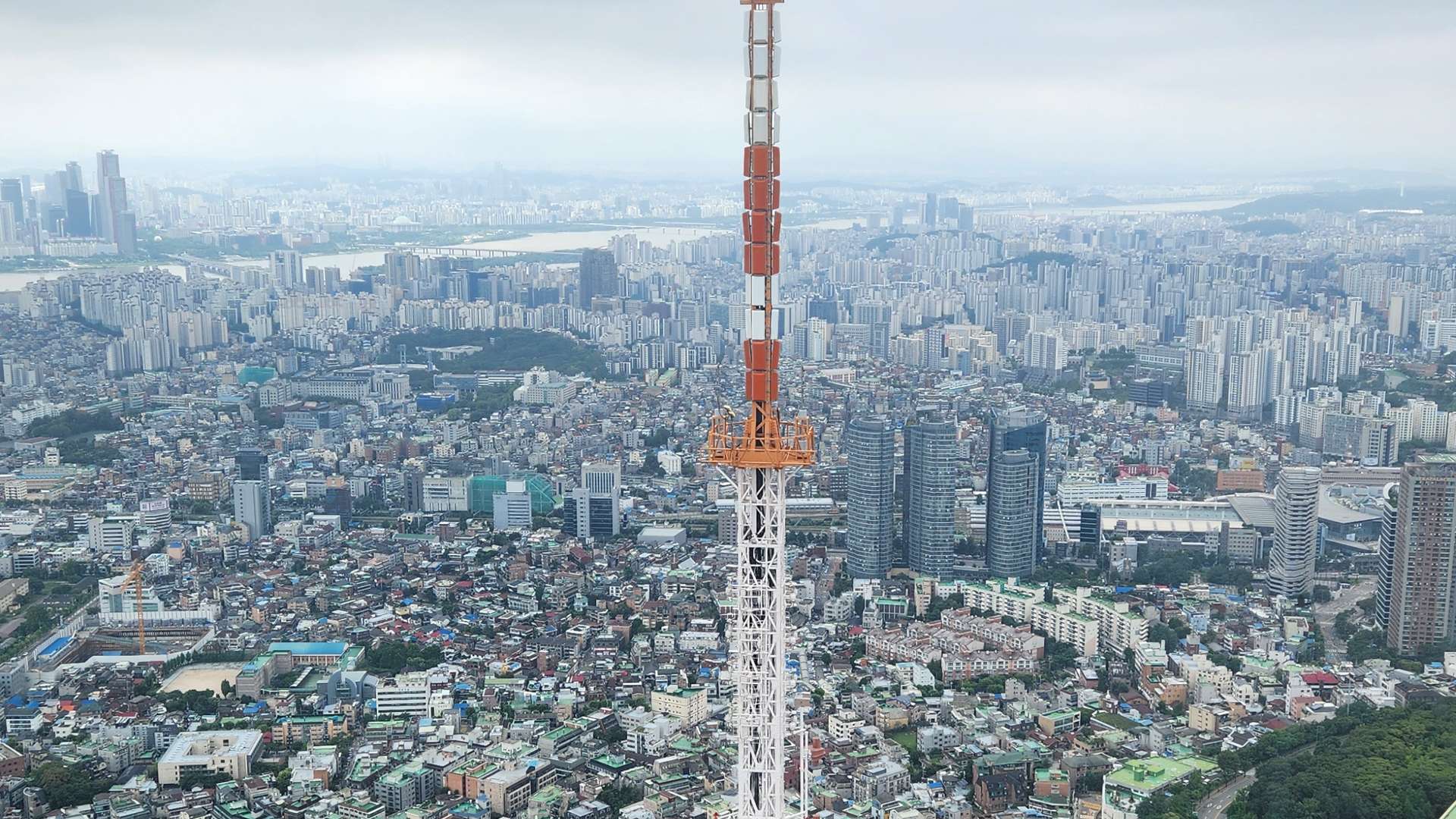 Tall orange and white broadcast tower rising above a dense cityscape under cloudy skies.