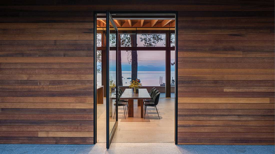 Modern dining area with glass pivot door framed by black metal, natural wood siding exterior, and panoramic lake views through floor-to-ceiling windows, featuring a rectangular wooden dining table with black chairs.