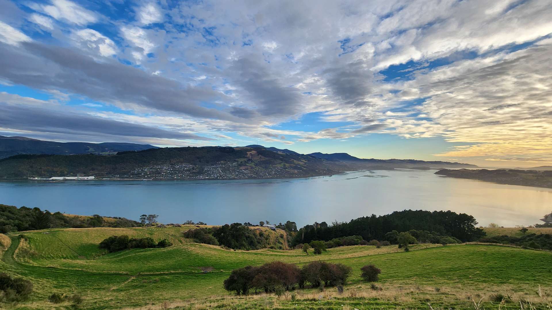 Wide view of a calm blue bay surrounded by green hills and distant mountains under a dramatic cloudy sky.