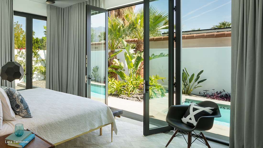 Modern bedroom with floor-to-ceiling glass doors opening to a private patio with pool, gray curtains, white bedding, and black accent chair.