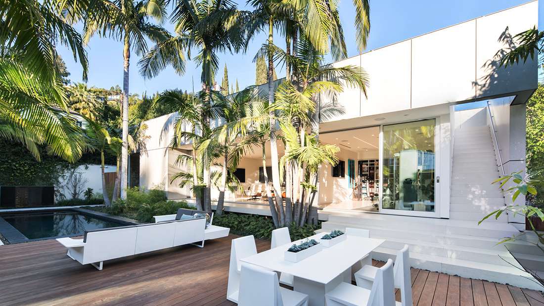 Modern outdoor living space with white minimalist furniture on a wooden deck, adjacent to a rectangular swimming pool, surrounded by tall palm trees and lush greenery, with large sliding glass doors opening to an open-concept interior.