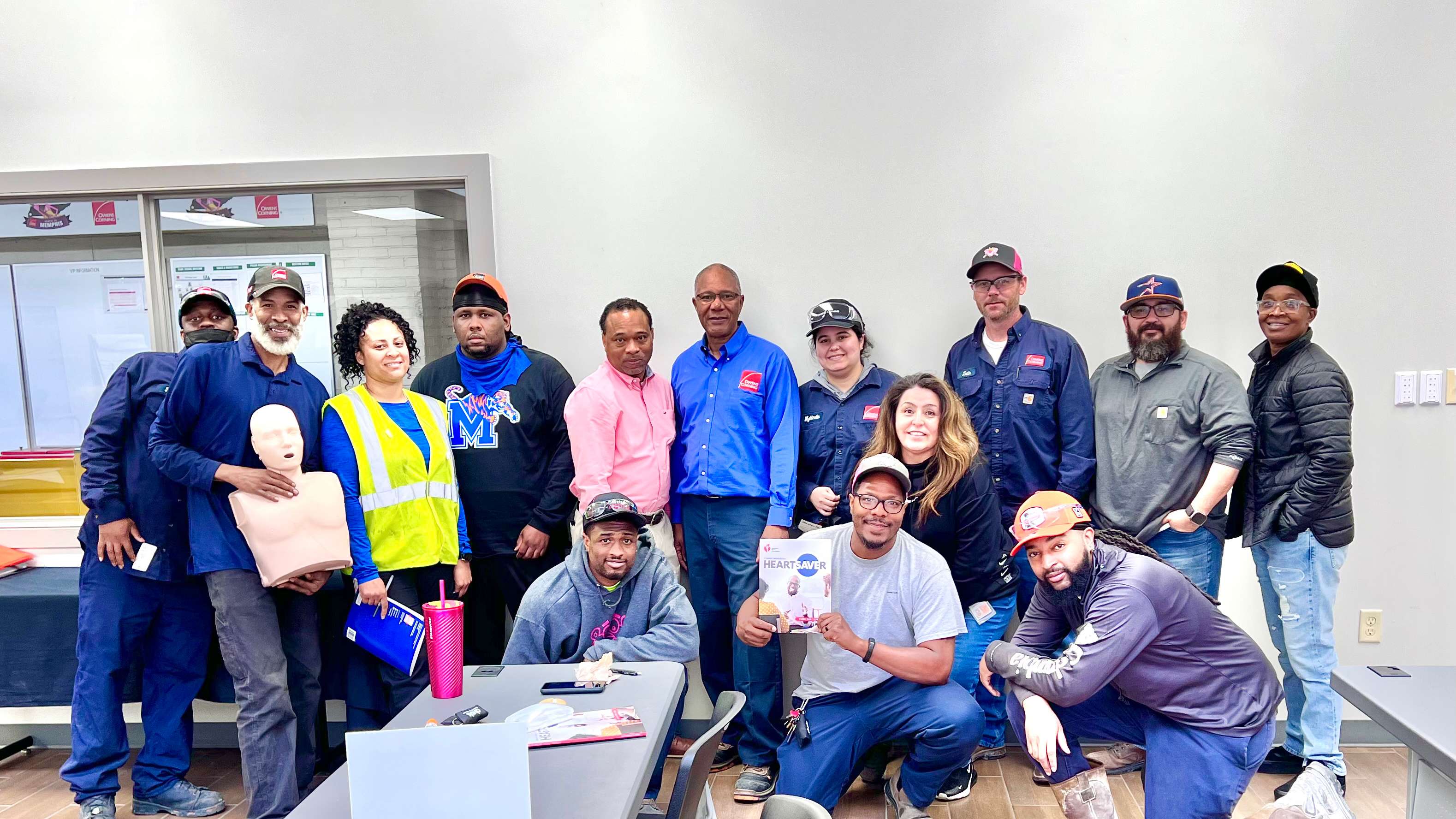 Group of workers in a training room posing together after a safety and CPR training session, with a CPR manikin and instructional materials visible on the table.