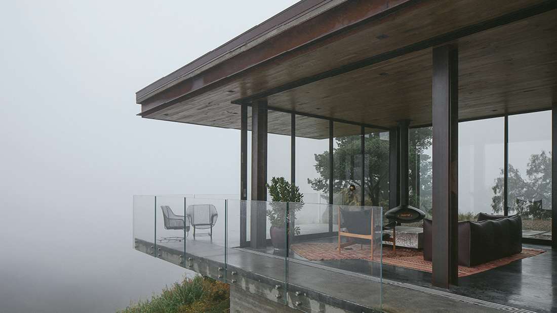 Modern cliffside home with cantilevered concrete deck, glass railing, floor-to-ceiling windows, and open-air lounge area featuring minimalist chairs and potted plants overlooking foggy mountain views.