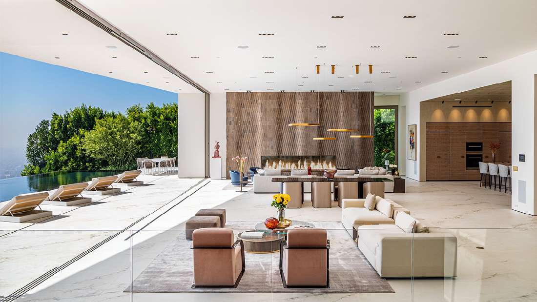  Luxury open-concept living space with marble flooring, modern lounge furniture, linear fireplace, and sliding glass walls opening to an infinity pool terrace with panoramic hillside views.
