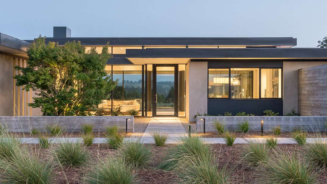 Contemporary single-story home with flat roof design, large glass entry door, black-framed windows, and minimalist landscaping featuring ornamental grasses and concrete planters.