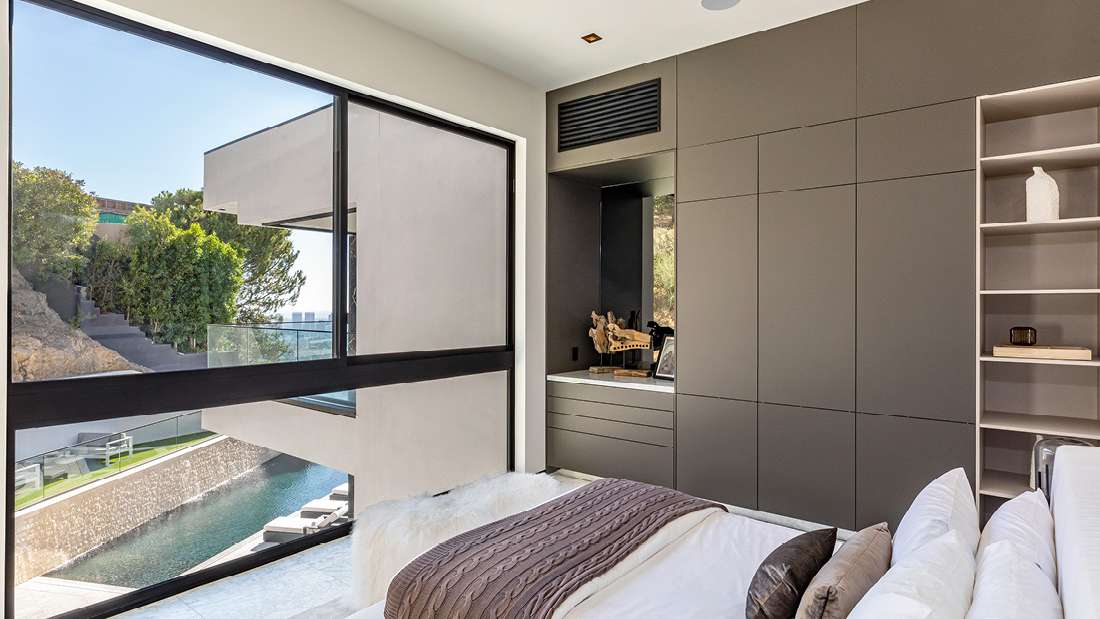 Luxury modern bedroom with large picture window overlooking hillside pool and contemporary architecture, featuring built-in matte gray cabinetry, open shelving, and minimalist decor.