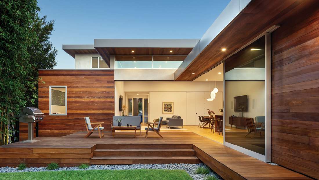 Contemporary home exterior with horizontal wood siding, large sliding glass doors, and elevated wooden deck featuring outdoor seating and built-in stainless steel grill.