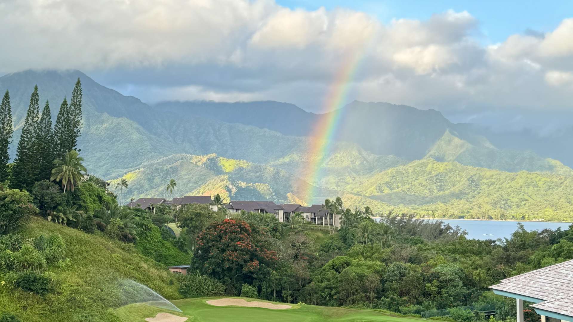 Rainbow over lush green valley with mountains and a golf course near a calm water pond.