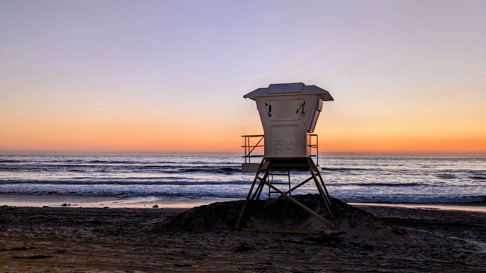 Lifeguard tower on a sandy beach at sunset with calm ocean waves and a colorful orange and purple sky.