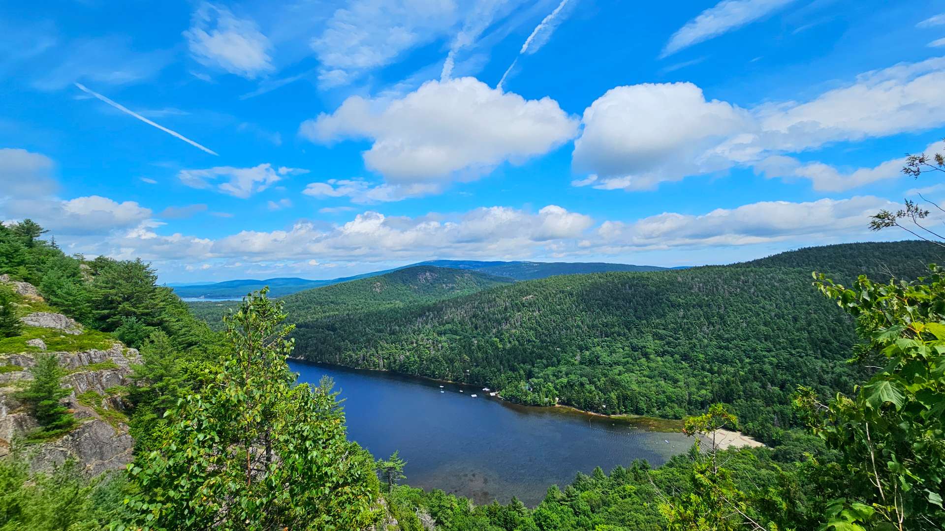 Scenic view of a forested mountain valley with a blue lake under a bright sky with scattered clouds.