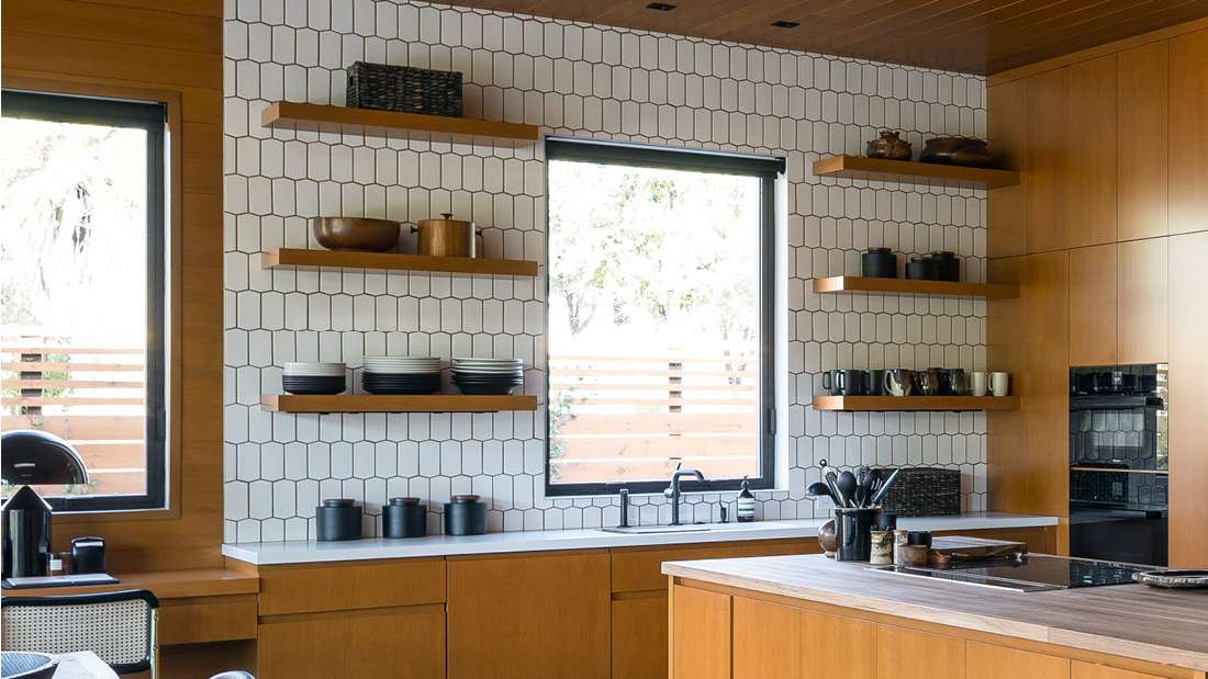 Modern kitchen with white hexagonal tile backsplash, floating wooden shelves displaying dishes and decor, large black-framed windows, and wood cabinetry with integrated appliances and island countertop.