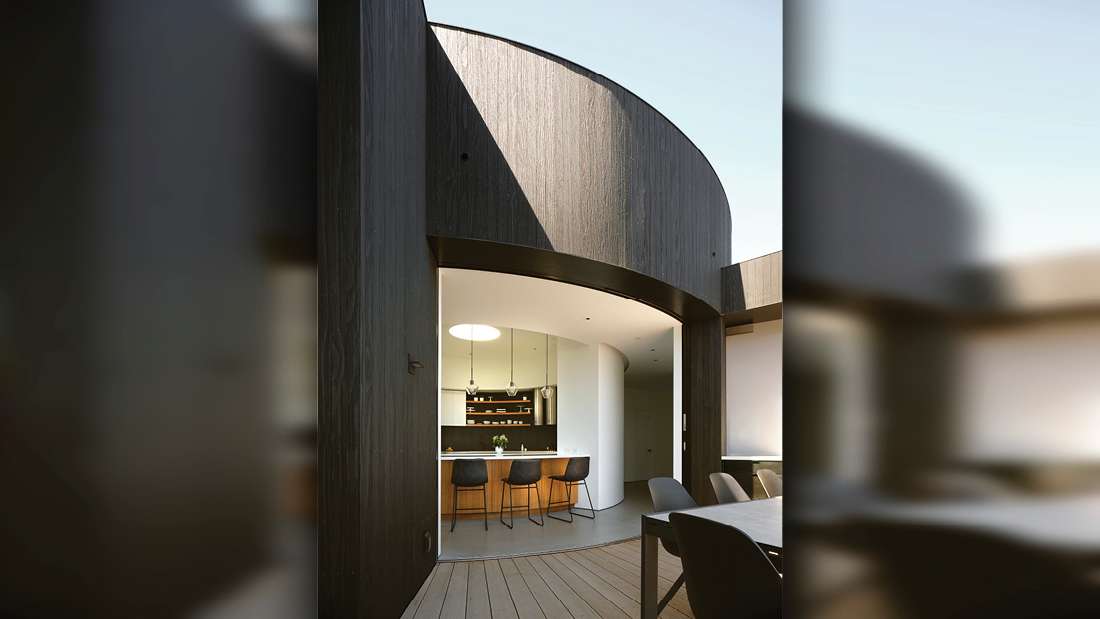 Contemporary home with curved architectural facade, dark wood cladding, and large glass doors opening to an outdoor deck with dining table and chairs.