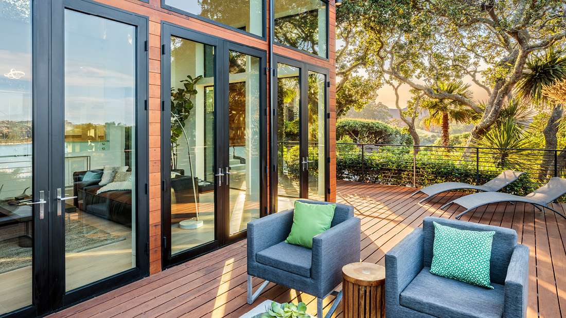 Modern outdoor deck with warm wood flooring, black-framed floor-to-ceiling glass doors, gray lounge chairs, and cushioned armchairs with green accent pillows overlooking lush garden and sunset views.