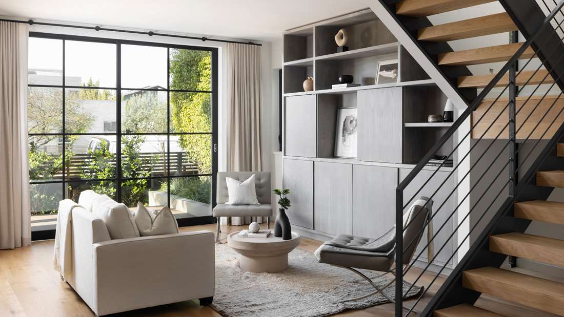 Bright modern living room with large grid-style black-framed windows, white sofa, gray accent chairs, round coffee table, built-in shelving, and open staircase with wooden steps and metal railing.
