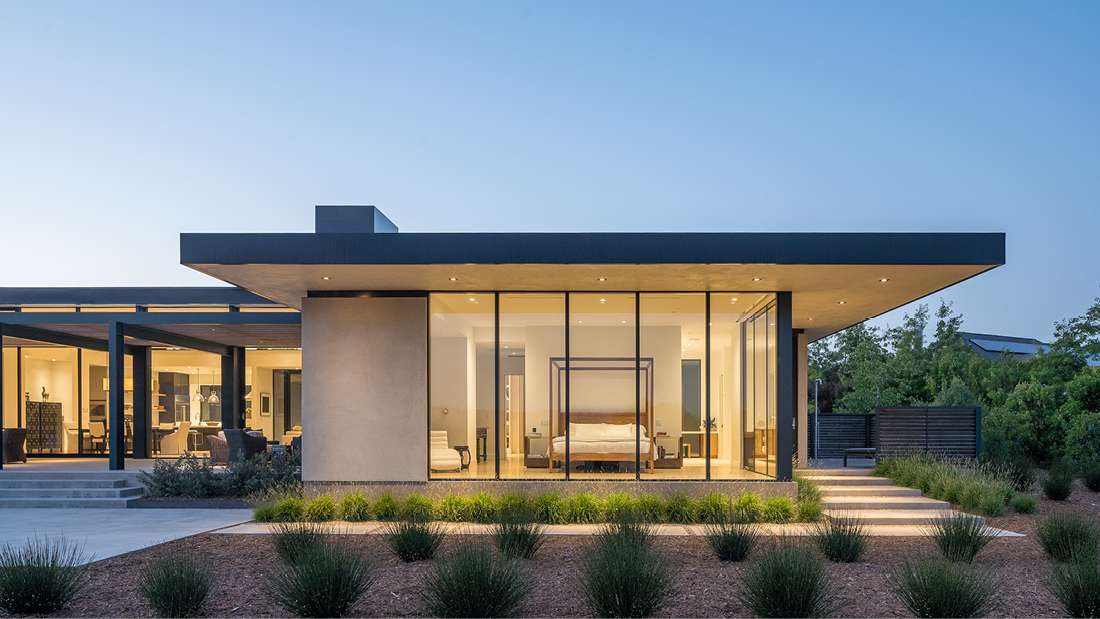Contemporary single-story home with flat roof design, floor-to-ceiling glass walls, minimalist landscaping featuring desert plants, and illuminated pathway leading to the entrance.