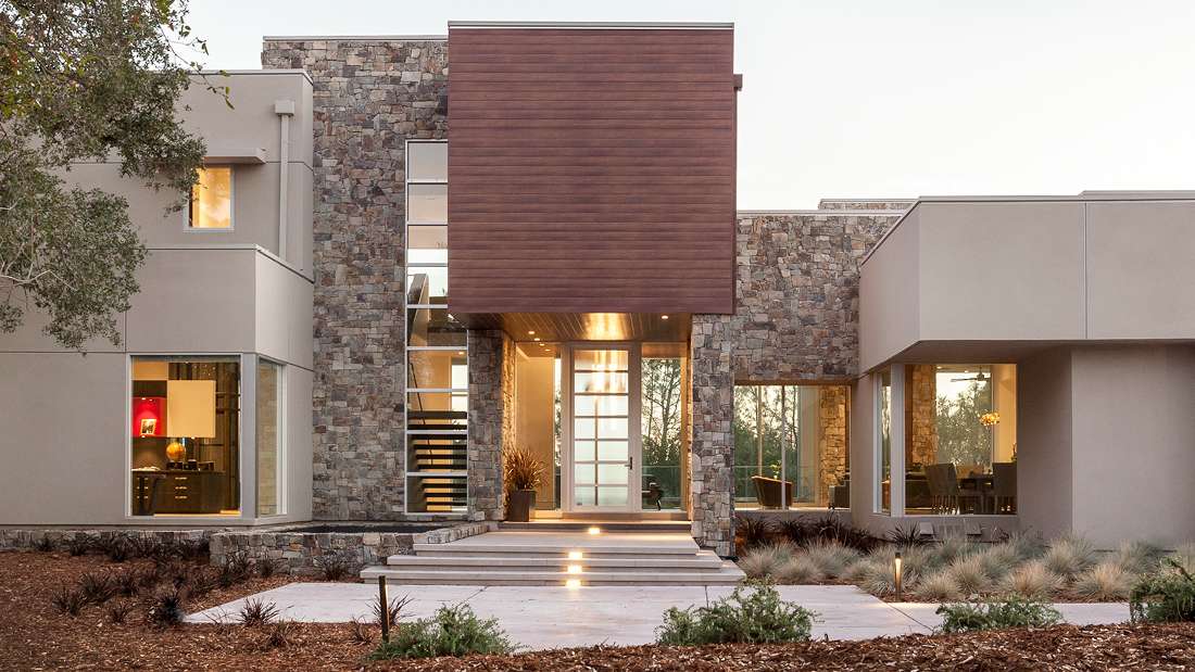 Contemporary luxury home exterior with stone cladding, large glass windows, and cantilevered wooden facade above a modern glass front door, surrounded by minimalist landscaping with ornamental grasses.