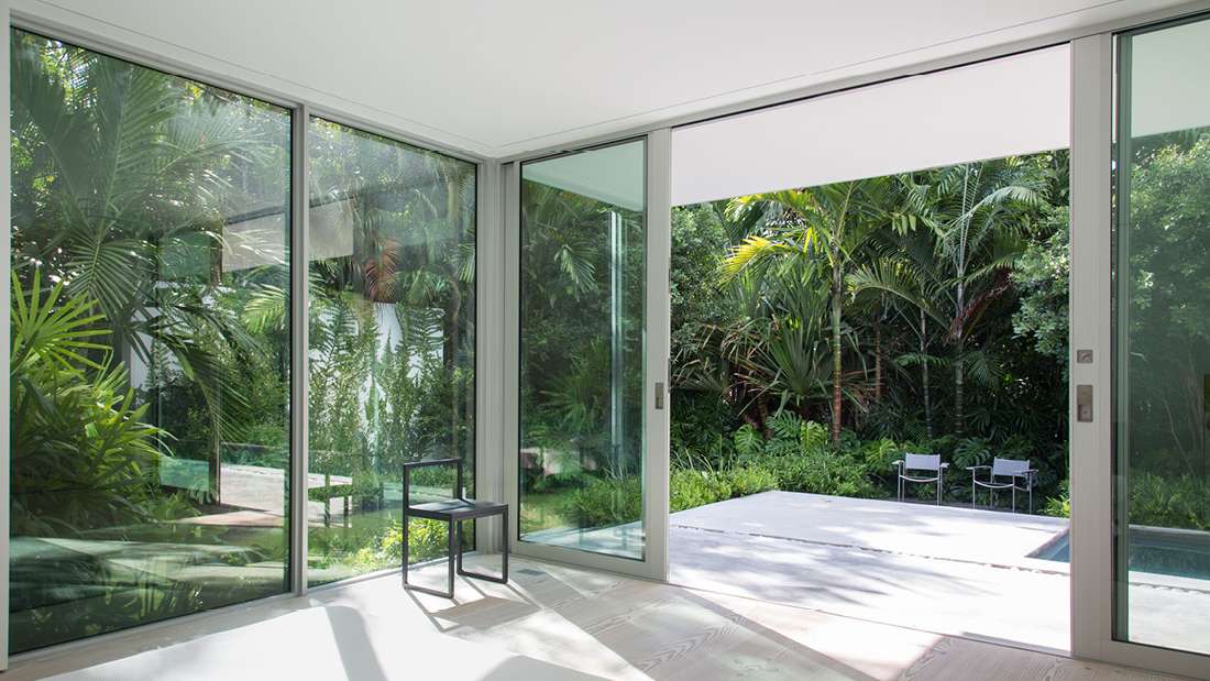 Minimalist indoor space with large sliding glass doors opening to a private patio and pool surrounded by lush tropical greenery, featuring light wood flooring and a single modern black chair.
