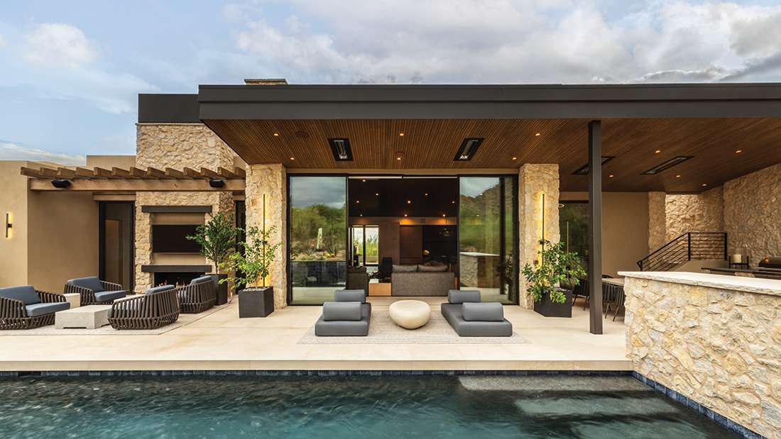 Contemporary luxury home with stone exterior, covered outdoor lounge, modern seating area, and private pool under a cloudy sky.