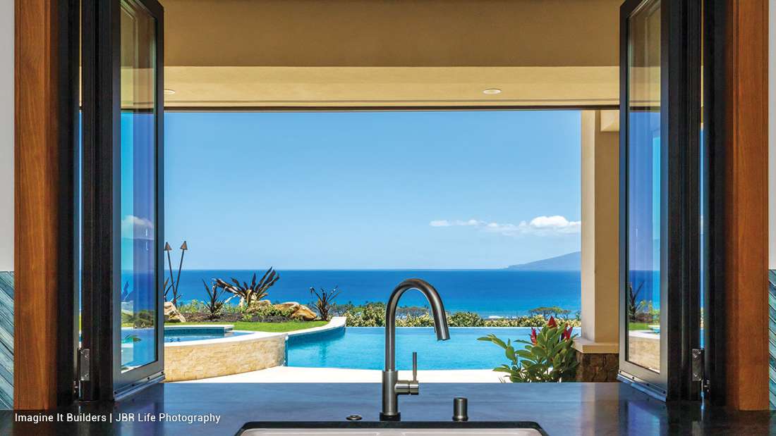 Luxury oceanfront kitchen view with stainless steel faucet and dark stone countertop, framed by open glass doors overlooking infinity pool, tropical landscaping, and panoramic blue ocean horizon.