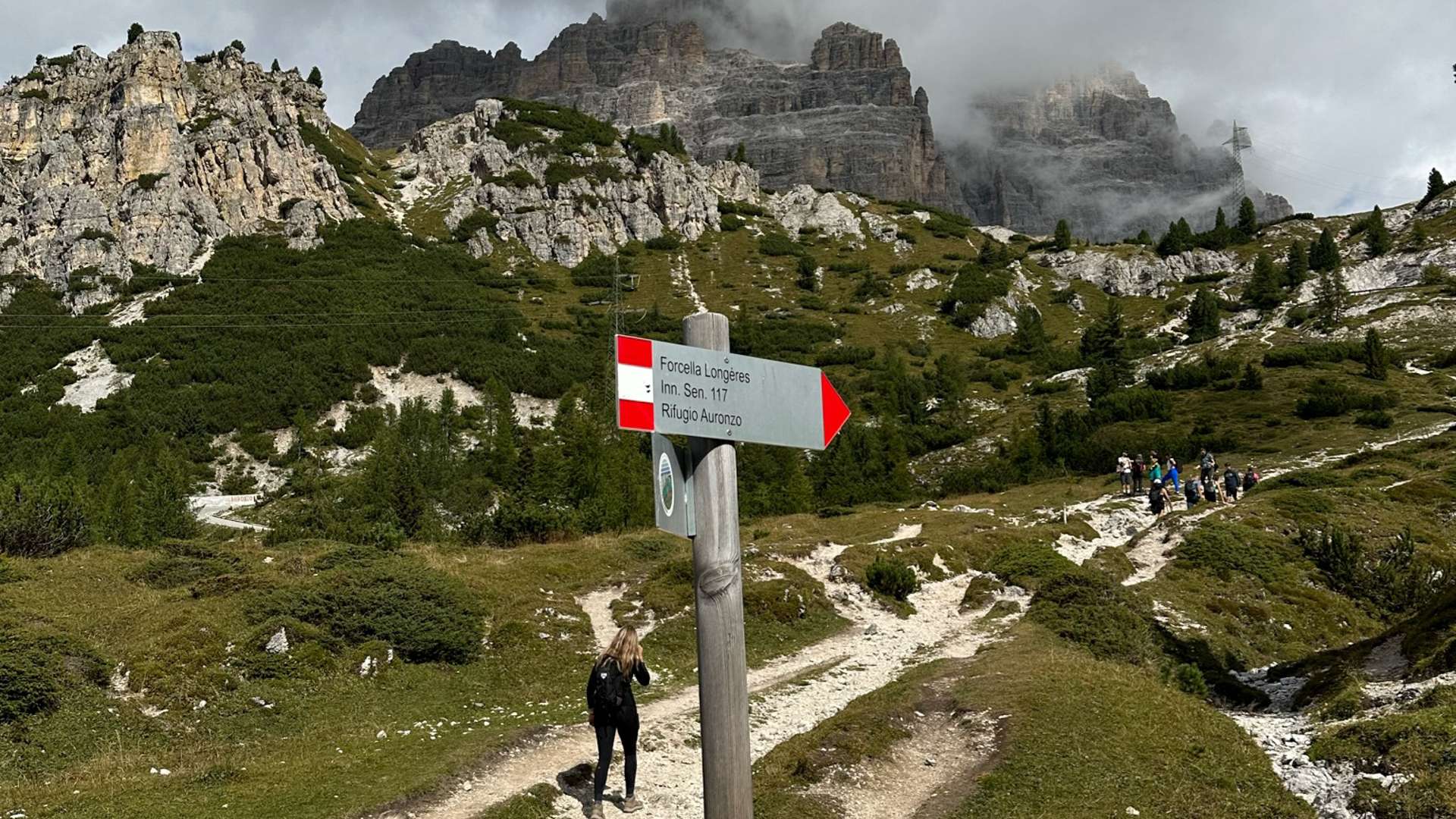  Mountain trail in the Dolomites with a wooden signpost and misty peaks under cloudy skies.