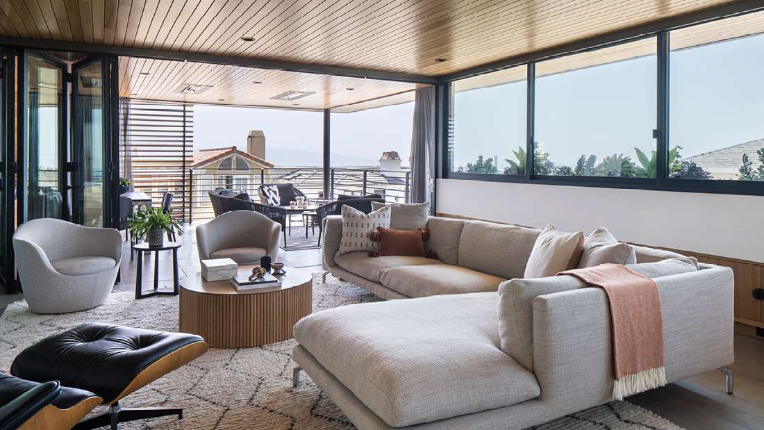 Modern living room with light gray sectional sofa, round wooden coffee table, sculptural lounge chairs, and folding glass doors opening to a covered balcony with outdoor seating and panoramic views.