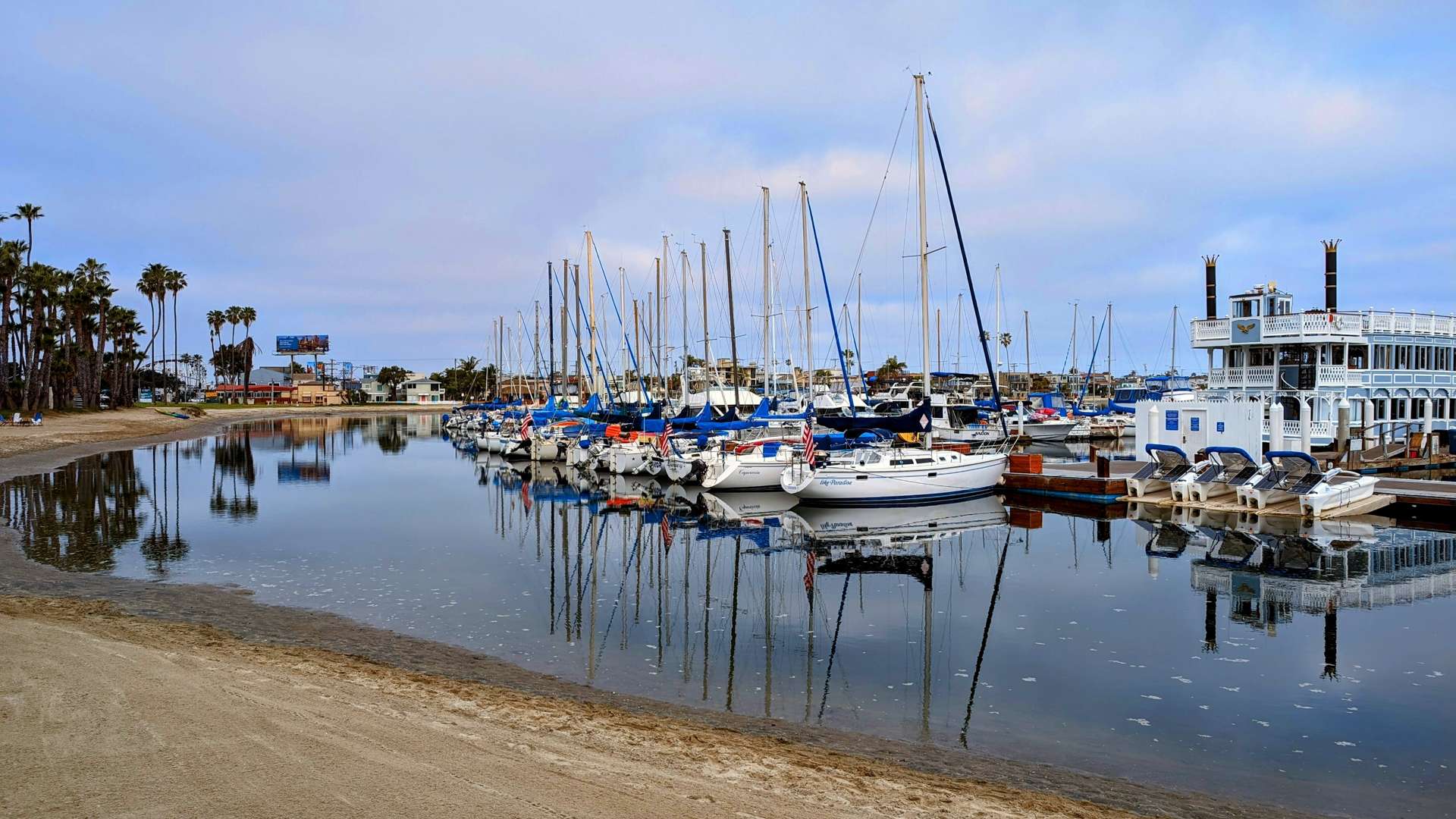 Row of sailboats docked in a calm marina with reflections on the water and palm trees in the background under a cloudy sky.