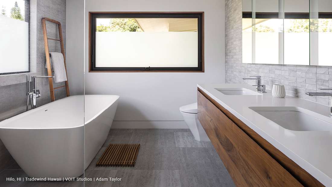 Modern bathroom with freestanding white bathtub, double-sink vanity featuring wood cabinetry and white countertop, large horizontal frosted window, and gray tile flooring.