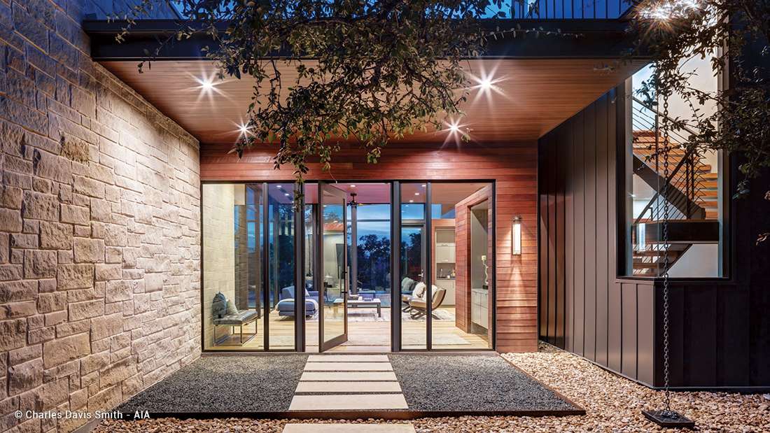 Modern home entryway with folding glass doors framed in black metal, natural stone wall on one side, vertical wood siding on the other, and covered porch with recessed lighting and minimalist landscaping.